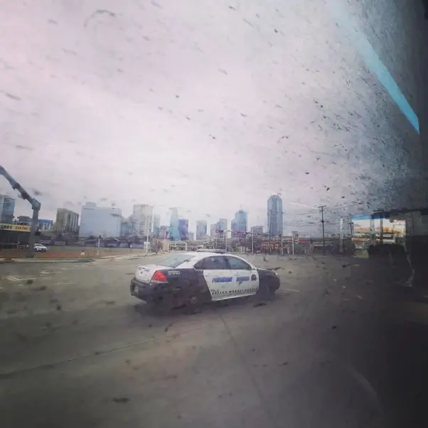 A police car is seen driving along a wide highway, viewed from a nearby dirt-covered bus window, with the skyline of Dallas in the background, on a cloudy, miserable day.