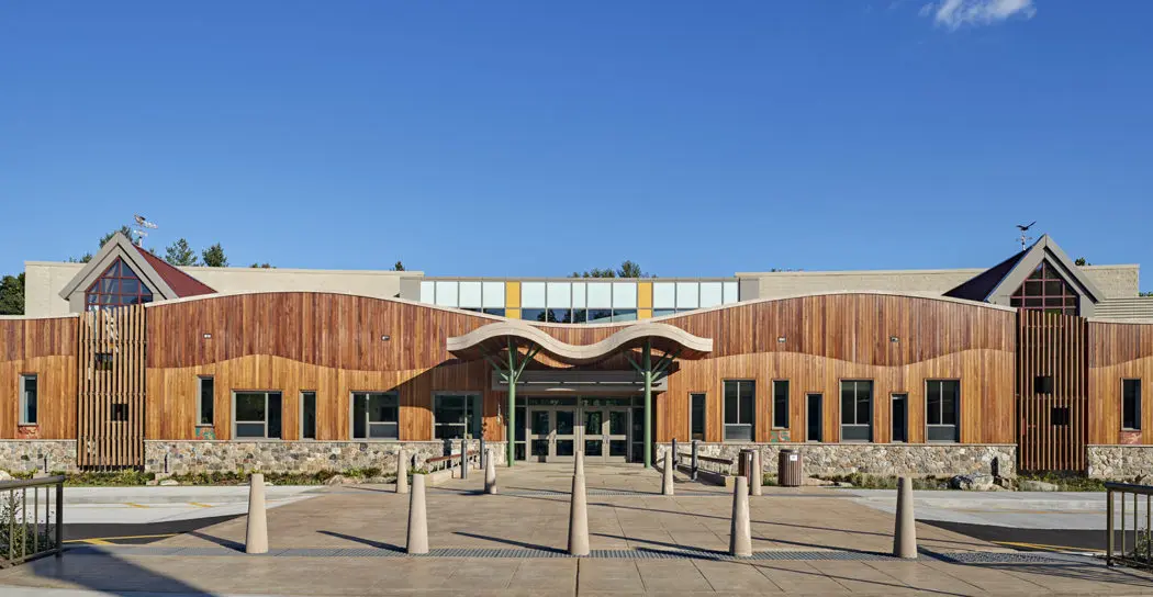 An exterior view of the school, with concrete pylons visible, as well as a wavy, rigid awning over the entrance. The façade of the building features darker wood, with a wavy boundary above the lower, lighter-coloured wood